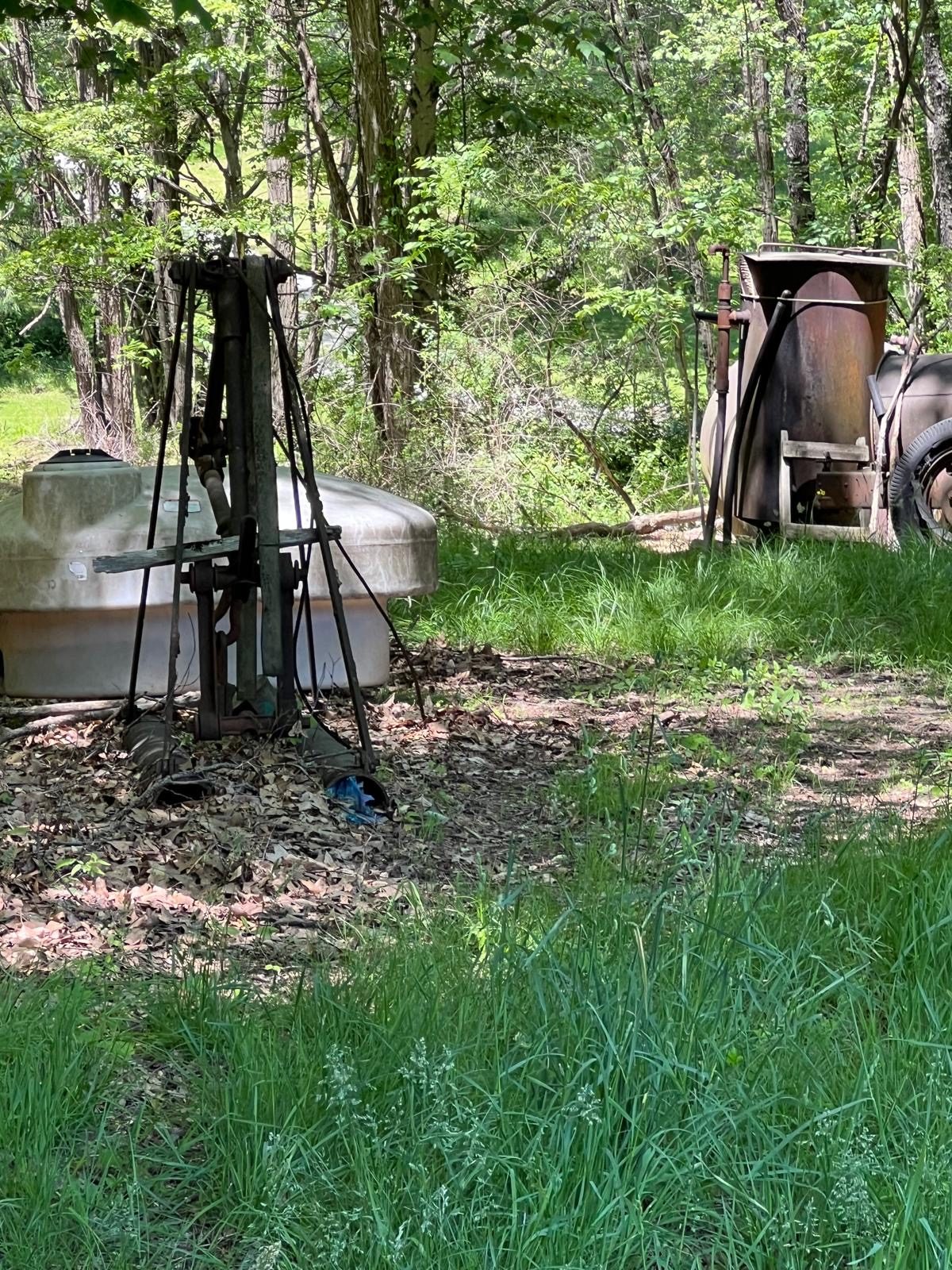 Una estructura metálica de trípode junto a un tanque de plástico blanco en un claro del bosque, con equipo oxidado en la línea de árboles.